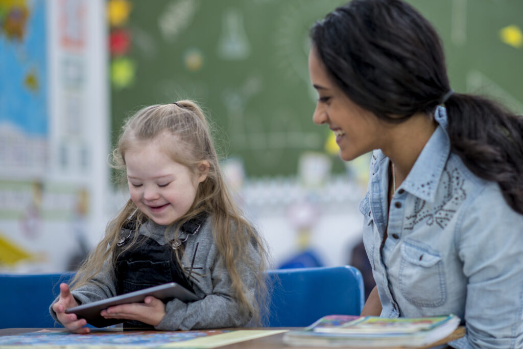 A girl with down syndrome is in a classroom with her teacher. She is happily playing with a tablet computer while being watched by her teacher.